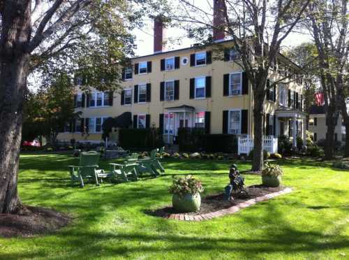 A large yellow house with black shutters, surrounded by green grass and trees, featuring outdoor seating and flower pots.