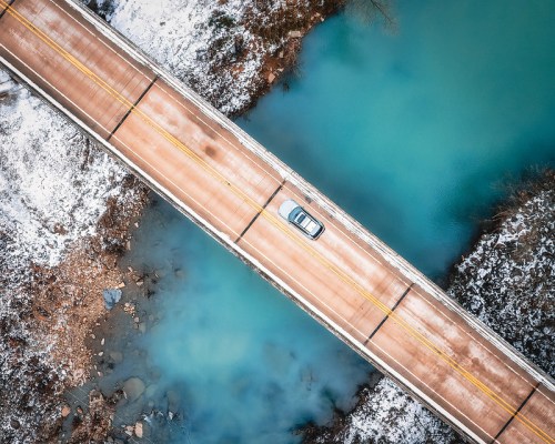 Aerial view of a car driving on a snowy bridge over turquoise water.