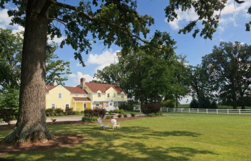 A yellow house with a red roof surrounded by trees and a green lawn, featuring two chairs in the foreground.