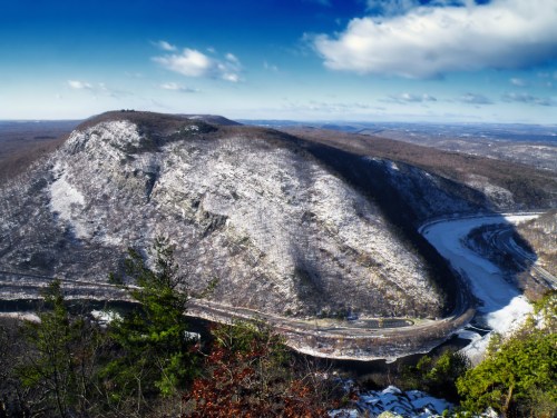 A panoramic view of a snow-covered mountain with a winding river below, under a clear blue sky with scattered clouds.