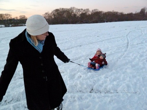 A woman pulls a child on a sled through a snowy field during sunset. Trees are visible in the background.