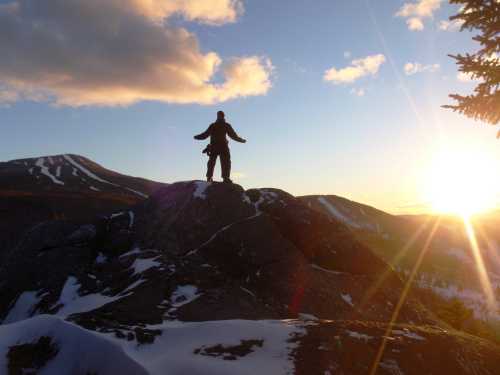 A silhouette of a person standing on a rock at sunset, with mountains and snow in the background.