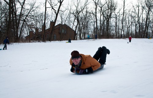 A person in an orange jacket sleds down a snowy hill, surrounded by trees and other people enjoying winter activities.
