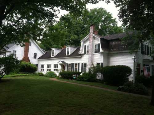 A charming white house with a sloped roof, surrounded by greenery and trees, featuring a small porch and an American flag.