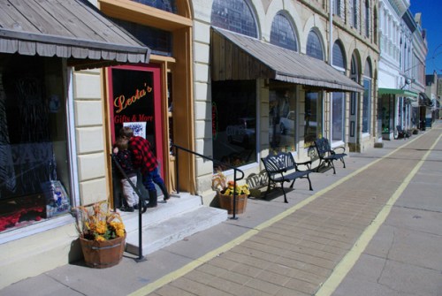 A child peeks into a shop entrance, with colorful flower pots and a sunny street lined with storefronts.