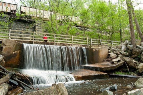A small waterfall cascades over a stone ledge, surrounded by greenery and rocks, with a train passing overhead.