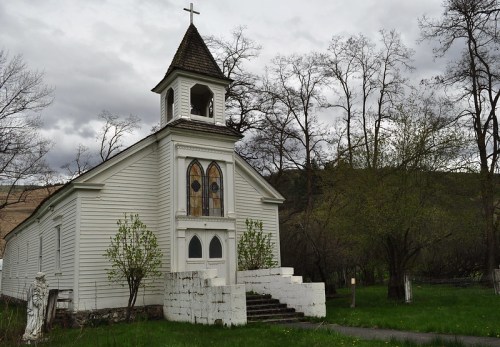 A small white church with a bell tower, surrounded by trees and a cloudy sky. Steps lead up to the entrance.