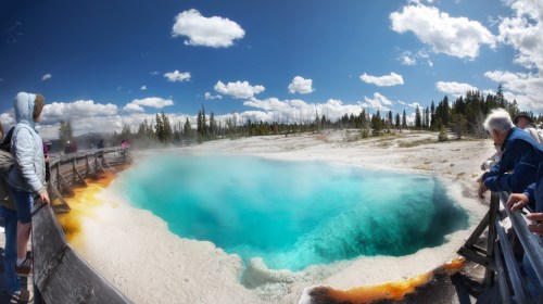 A vibrant blue hot spring surrounded by wooden railings and lush greenery under a partly cloudy sky.