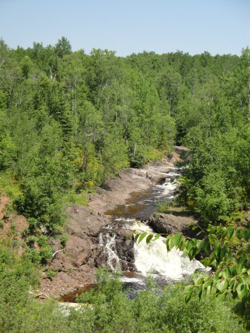 A serene river flows through a lush green forest, with rocky banks and gentle waterfalls under a clear blue sky.