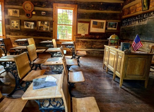 A rustic classroom with wooden desks, a teacher's desk, a globe, and an American flag, featuring a chalkboard and historical maps.
