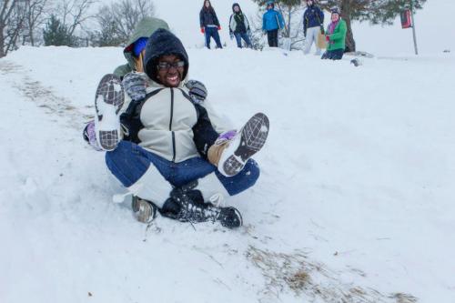Two children joyfully sledding down a snowy hill, with others playing in the background.