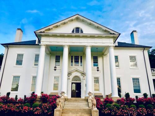 A grand white mansion with columns, surrounded by colorful flowers and a blue sky.