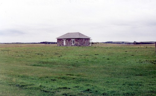 A solitary stone house stands in a vast green field under a cloudy sky.