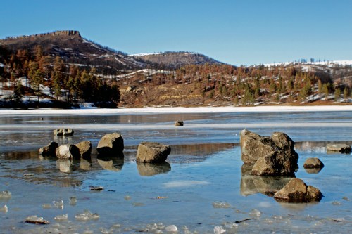 A serene frozen lake with scattered rocks, surrounded by snow-covered hills and clear blue skies.