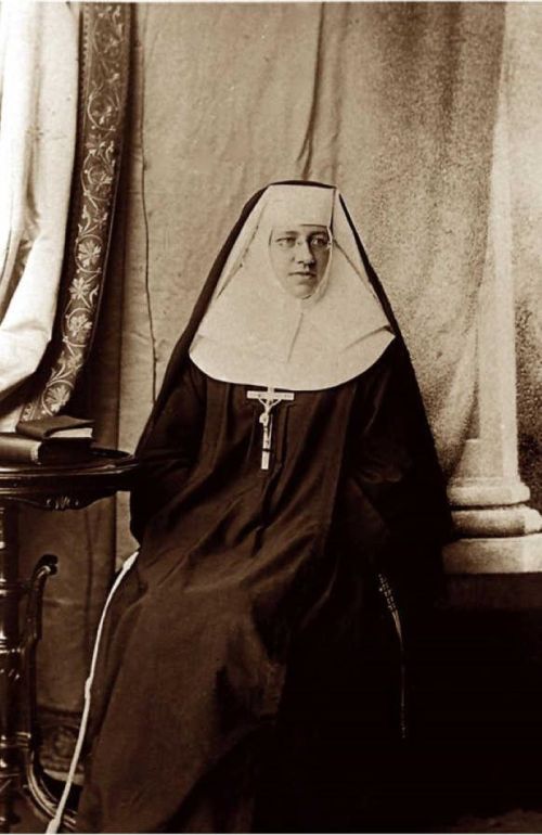 A historical black-and-white portrait of a nun seated, wearing traditional religious attire and a cross necklace.