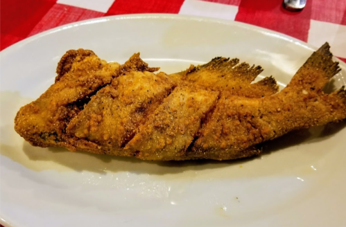 A plate with a crispy, fried whole fish, garnished and served on a white dish against a red and white checkered tablecloth.