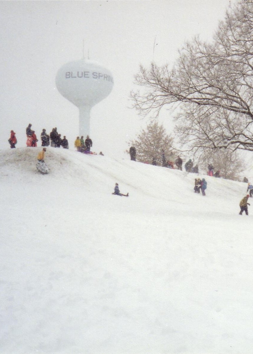 A snowy hill with people sledding, a water tower labeled "BLUE SPRING" in the background, and trees nearby.