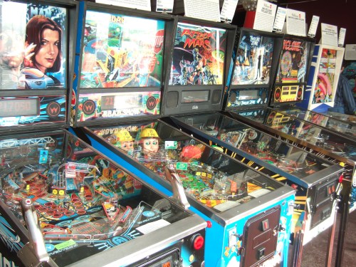 A row of colorful pinball machines with various themes and artwork, showcasing their playfields and controls.
