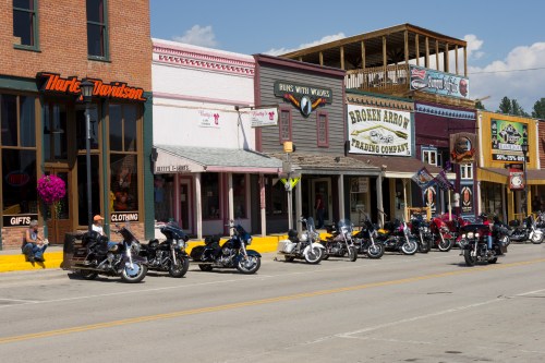 A row of motorcycles parked in front of colorful storefronts in a small town. Bright blue sky overhead.