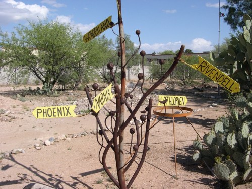 A whimsical signpost with yellow arrows pointing to fictional places like Wonderland, Rivendell, and Narnia in a desert setting.