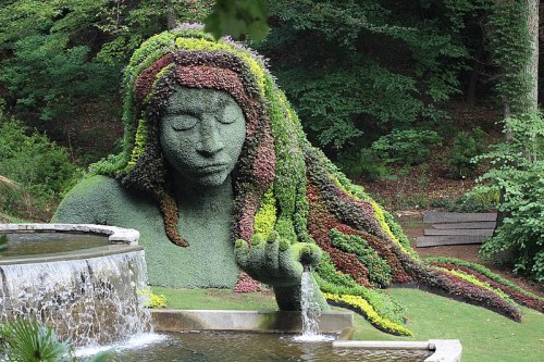 A large, green sculpture of a woman with flowing hair made of plants, set beside a waterfall in a lush garden.