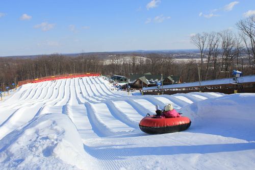 A snowy tubing hill with multiple lanes, people enjoying the ride, and a lodge in the background under a clear blue sky.