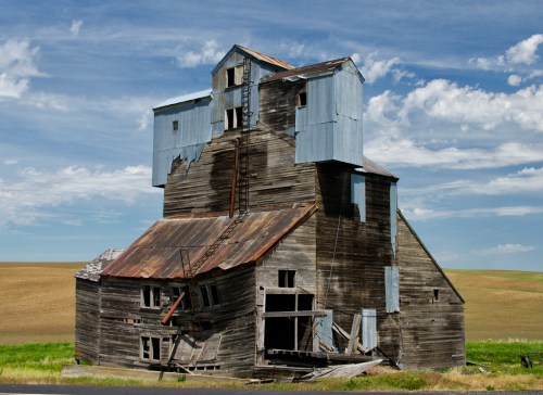 A weathered, multi-level barn with a blue metal roof, showing signs of decay against a backdrop of fields and blue sky.