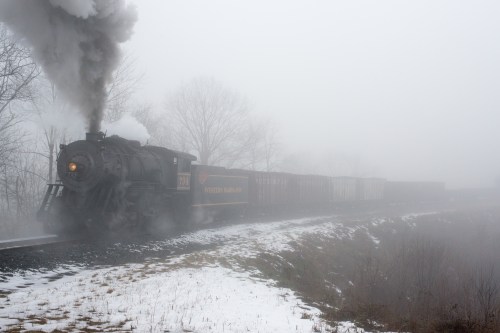 A steam locomotive chugs through a foggy landscape, with smoke billowing and snow on the ground.
