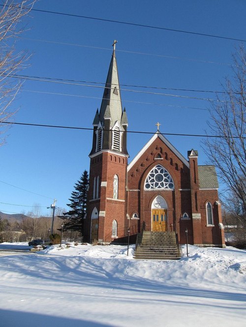 A brick church with a tall steeple, surrounded by snow and trees, under a clear blue sky.