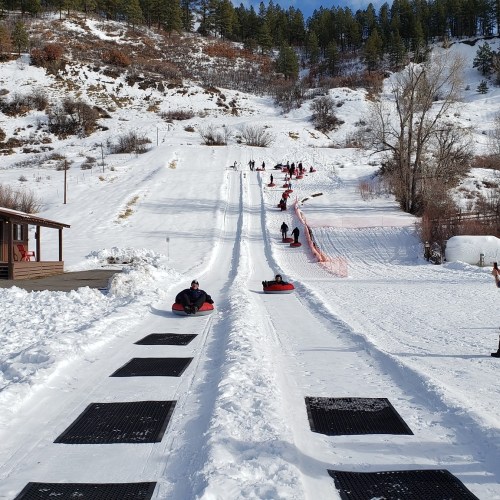 People tubing down a snowy hill, with trees in the background and a clear blue sky above.