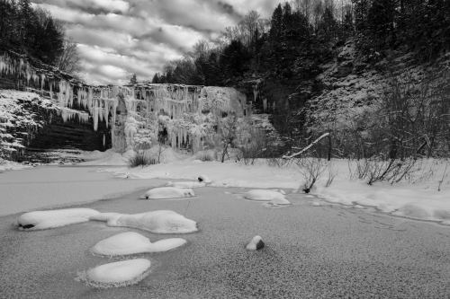 A frozen waterfall surrounded by snow-covered trees and icy terrain, captured in black and white.