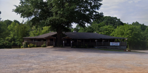 A rustic building surrounded by trees, with a gravel parking area and a sign at the entrance.