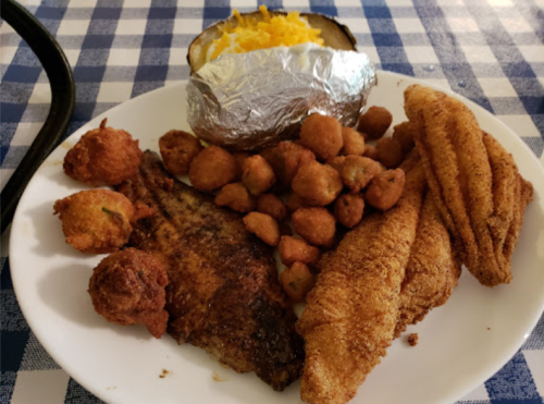 A plate of fried fish, hushpuppies, fried okra, and a baked potato topped with cheese on a checkered tablecloth.