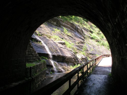 A view through a stone tunnel, revealing a rocky landscape with a small waterfall and a wooden path beside a stream.