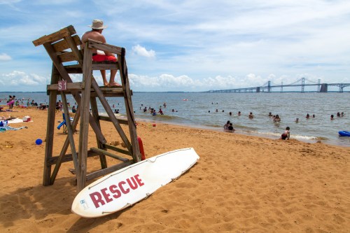 A lifeguard sits on a tall chair overlooking a sandy beach with people swimming and a bridge in the background.