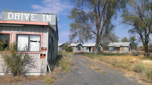 Abandoned drive-in building with weathered signage, surrounded by overgrown grass and dilapidated structures in the background.