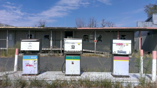 Abandoned gas station with old fuel pumps in front of a dilapidated building and overgrown weeds.