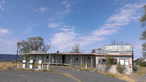 Abandoned drive-in theater with a weathered building and overgrown grass under a blue sky.