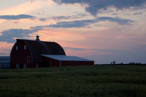 A red barn stands in a field at sunset, with colorful clouds in the sky and a peaceful rural landscape.