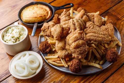 A large platter of fried catfish, French fries, hushpuppies, coleslaw, and sliced onions on a wooden table.