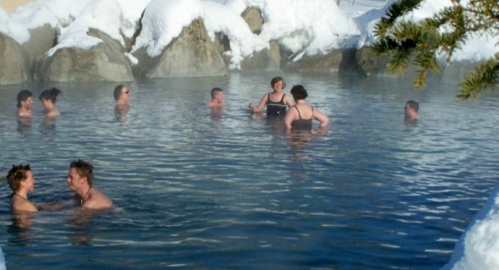 People enjoying a hot spring surrounded by snow-covered rocks and trees on a sunny winter day.