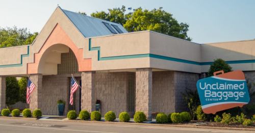 Exterior of the Unclaimed Baggage Center, featuring a colorful sign and American flags, surrounded by greenery.