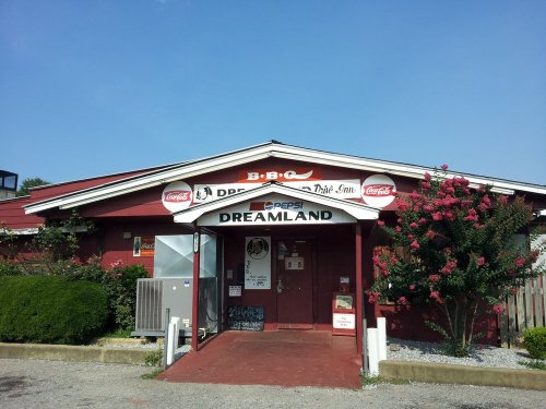 Exterior of Dreamland BBQ restaurant with a red facade, signage, and greenery in front under a clear blue sky.