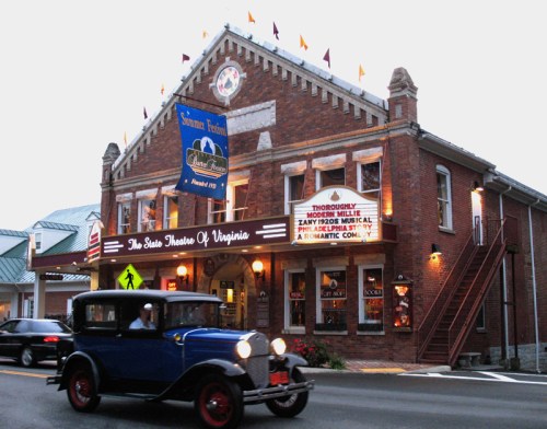 A vintage blue car drives past a historic brick theater adorned with flags and a marquee in Virginia.