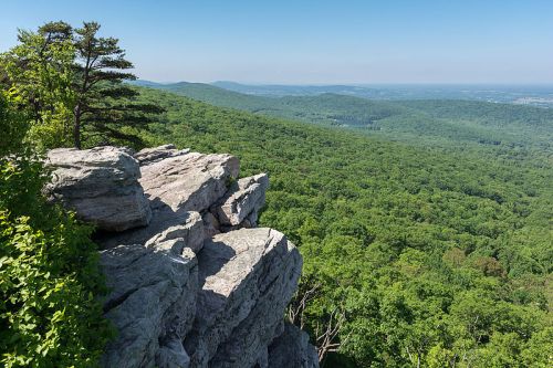 A rocky outcrop overlooks a lush green valley and distant mountains under a clear blue sky.