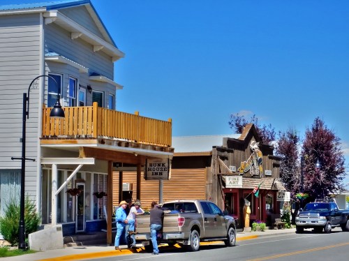 A sunny street scene featuring a rustic inn, art shop, and people unloading a truck in a small town.