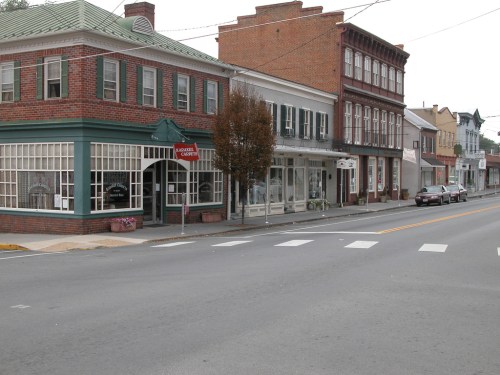 A quiet street scene featuring brick buildings, shops, and a tree, with a car parked nearby.
