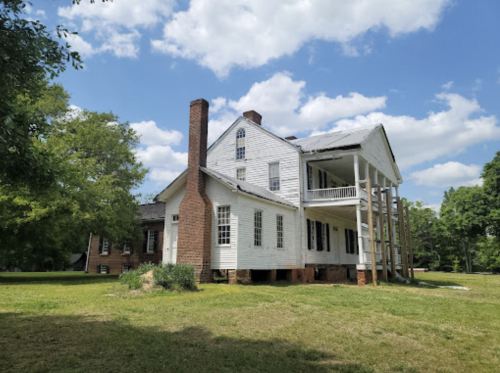 A historic white house with a porch, tall chimney, and green lawn under a blue sky with fluffy clouds.
