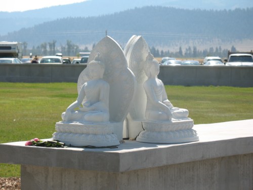 Two white Buddha statues seated on a stone platform, surrounded by green grass and mountains in the background.