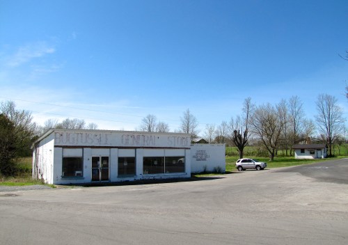 A small, white general store with faded signage, surrounded by trees and a clear blue sky. A car is parked nearby.